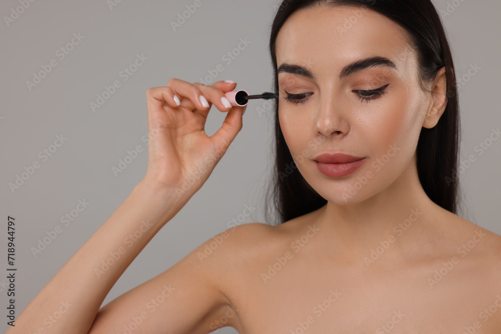 Beautiful young woman applying mascara on grey background