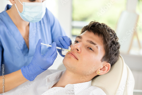 Closeup face of young male client receiving injections during lip enhancement procedure, professional cosmetologist hands in rubber gloves holding syringe