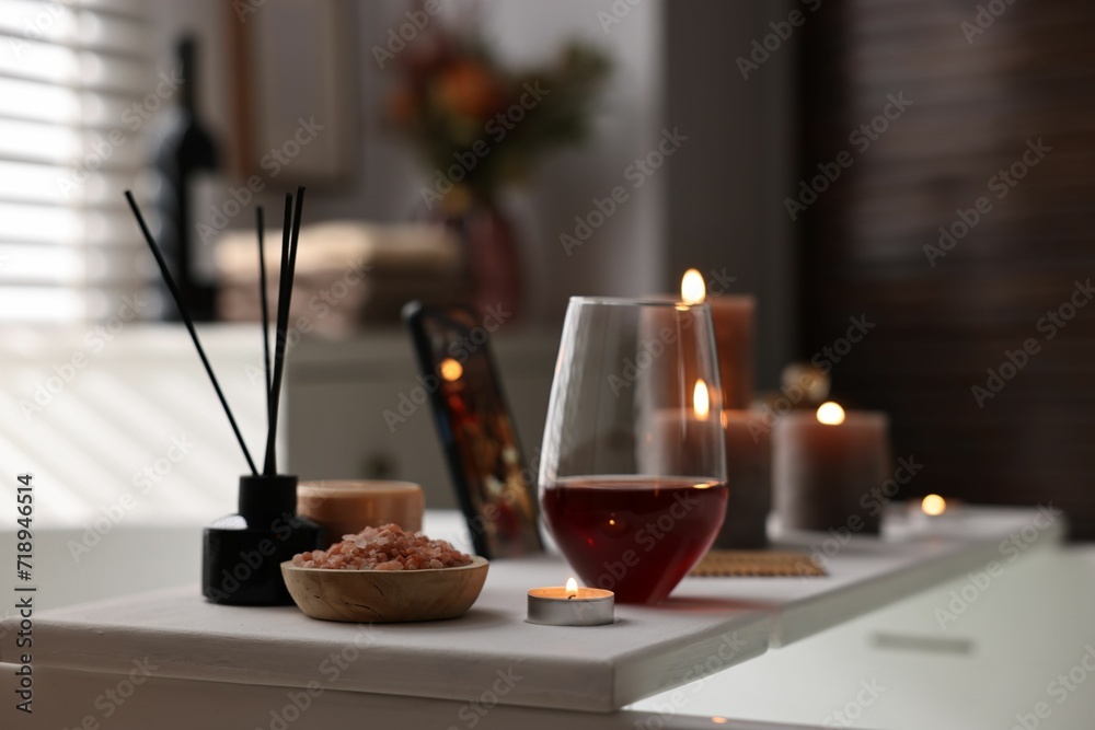 White wooden tray with glass of wine, beauty products and burning candle on bathtub in bathroom