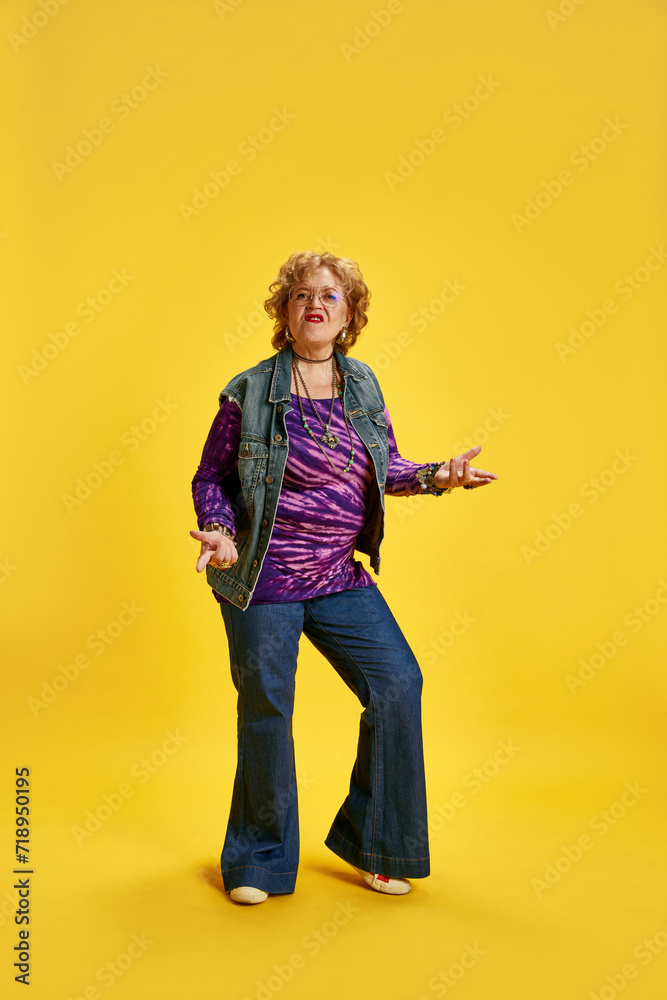 Elderly emotional woman in denim jeans purple top dancing against vibrant yellow background. Concept of empowerment of older adults, active seniors in modern life, fashion and style. Elderly emotional woman in denim jeans purple top dancing against vibrant yellow background. Concept of empowerment of older adults, active seniors in modern life, fashion and style.