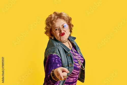 Senior woman puckering lips in round glasses dressed denim jacket  purple T-shirt with gold jewelry against yellow background. Concept of empowerment of older adults  active seniors in modern life.