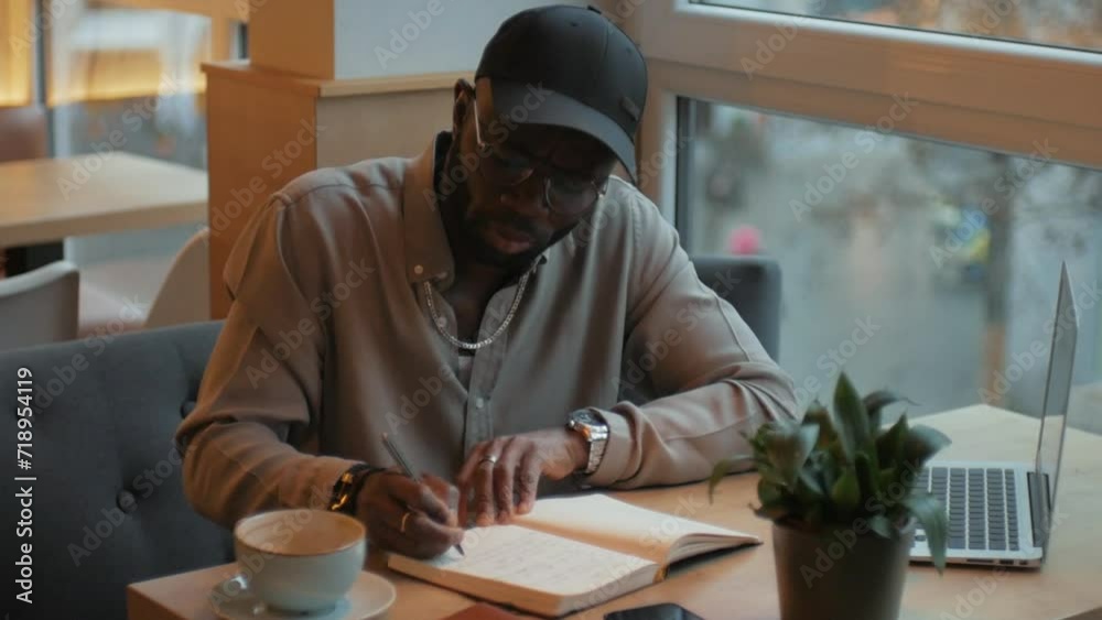 An African American man is writing in a notebook. Thinks about ideas. Sitting at a table with a laptop. Camera movement An African American man is writing in a notebook. Thinks about ideas. Sitting at a table with a laptop. Camera movement