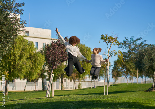 two sisters with curly afro hair have fun playing jumping on the grass in a city park on a sunny day