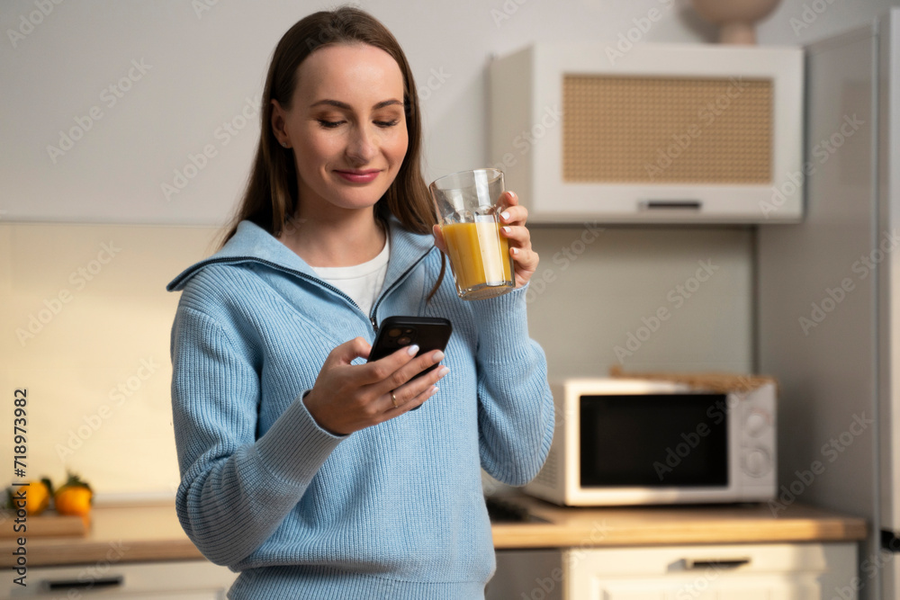 Young smiling happy woman in a blue sweater, using a mobile phone, drinking orange juice in the bright kitchen at home. 