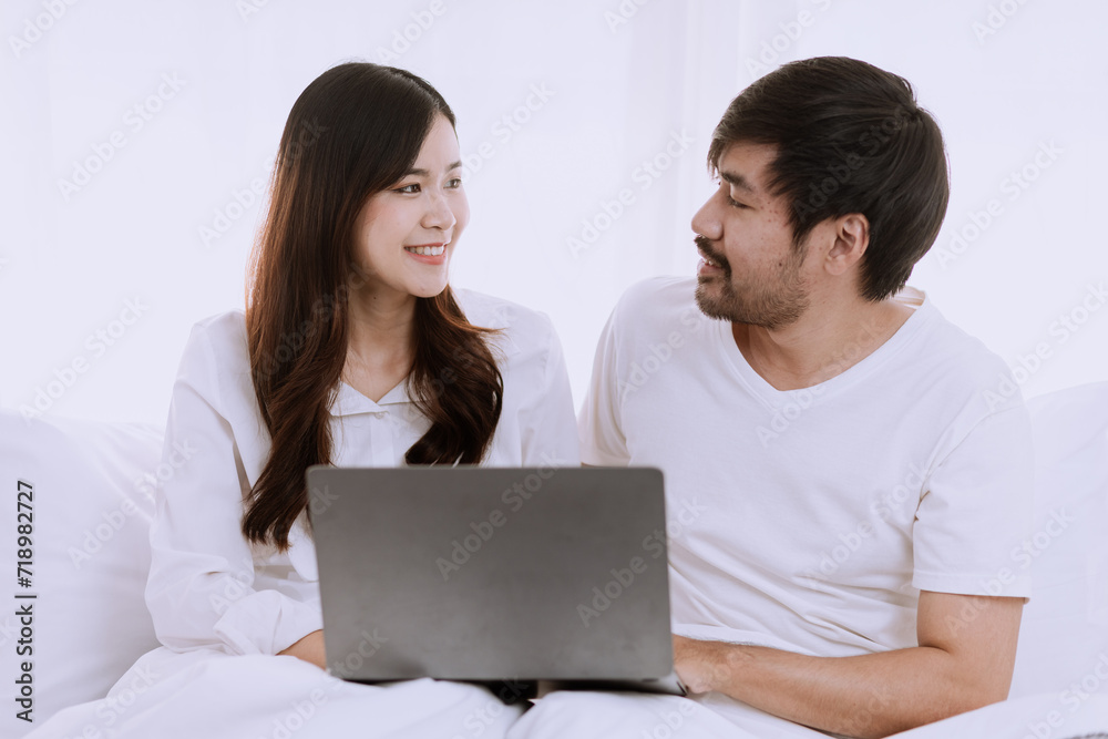 Happy asian man and woman using laptop sitting relaxed on a bed at home enjoy, Happy lover hug each other and smiling on the bed, Male and female couple concept.