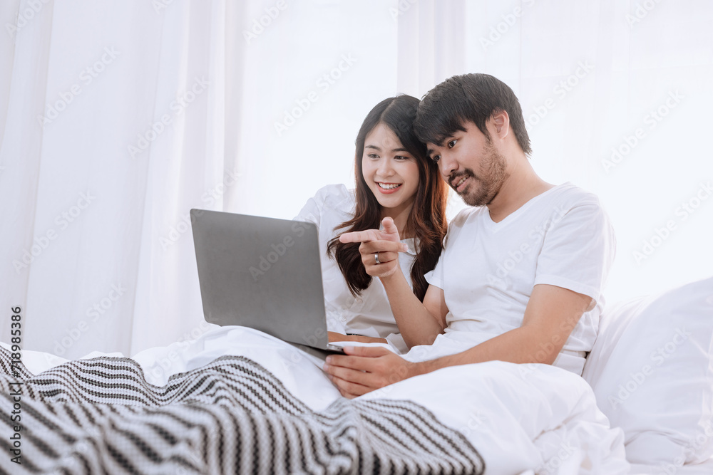 Happy asian man and woman using laptop sitting relaxed on a bed at home enjoy, Happy lover hug each other and smiling on the bed, Male and female couple concept.