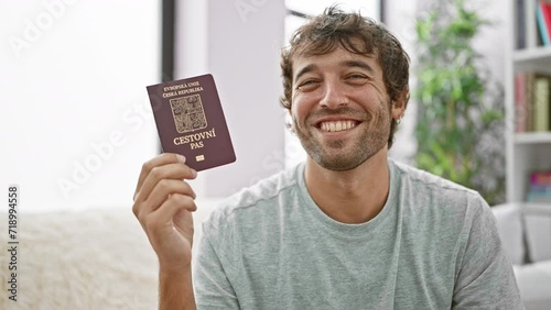 Confident bearded young man sitting on his living room sofa, grinning wide with czech republic passport, overflowing with happiness, ready to embark on an exciting vacation photo