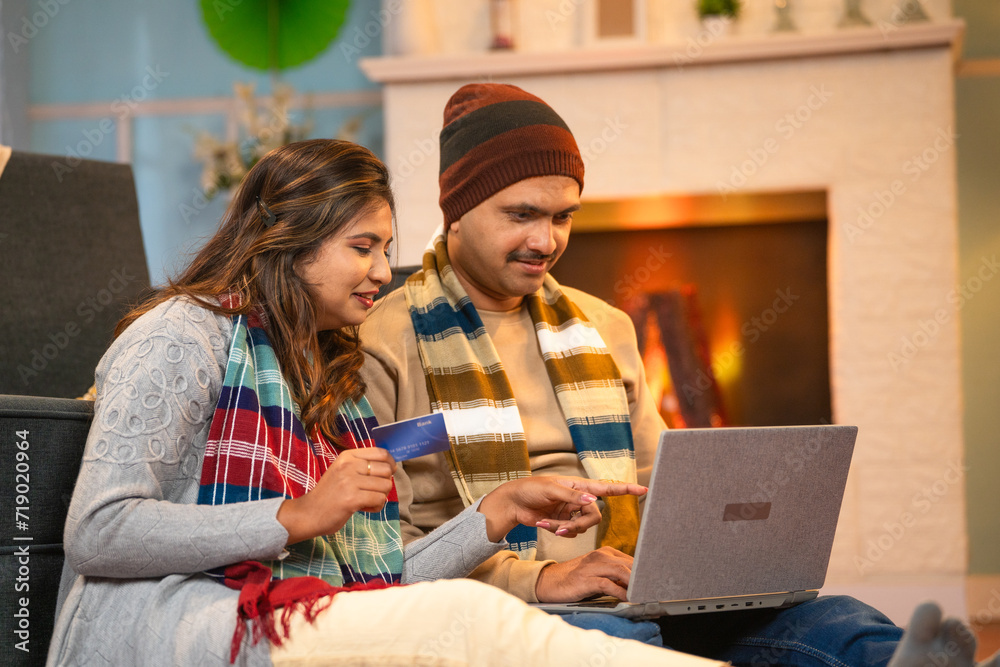 joyful Indian couples in winter wear doing ecommerce shopping on laptop using credit card at home - concept of online order, technology and banking or financial joyful Indian couples in winter wear doing ecommerce shopping on laptop using credit card at home - concept of online order, technology and banking or financial