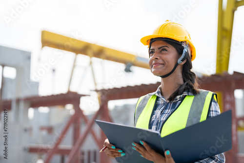 Asian woman engineer holding document smiling at construction site. Confident female Indian wearing protective helmet and vest working in factory making precast concrete wall for real estate housing. photo