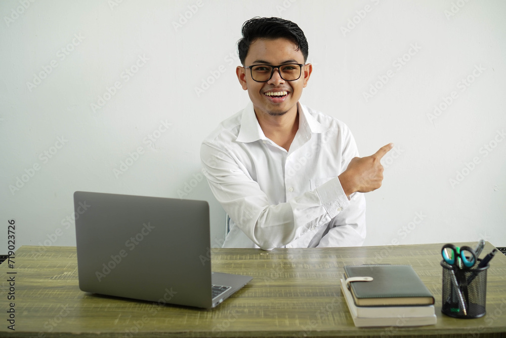 young asian businessman in a workplace smiling with pointing back, wearing white shirt with glasses isolated young asian businessman in a workplace smiling with pointing back, wearing white shirt with glasses isolated