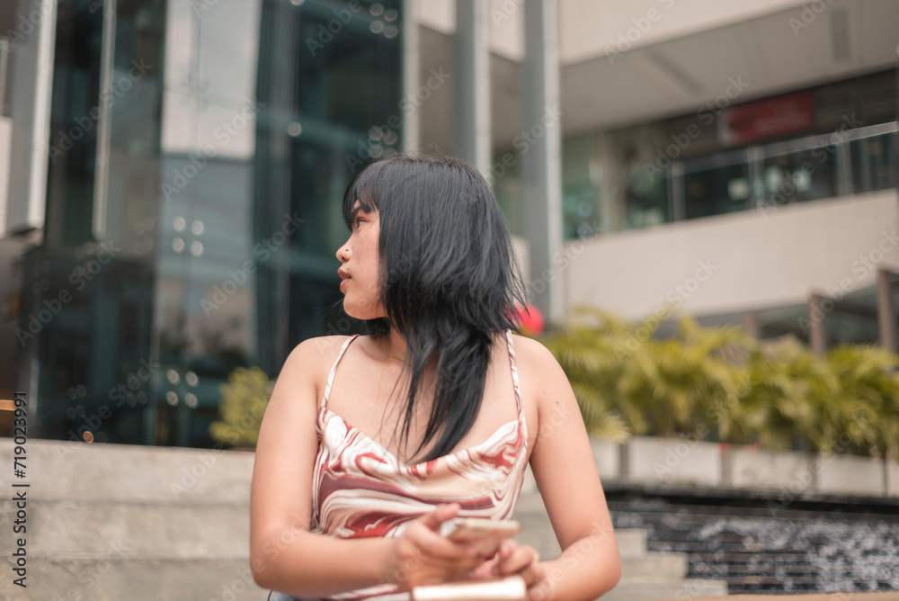 A thoughtful young Asian lady in casual attire looks away while sitting on a bench outside a modern building. A thoughtful young Asian lady in casual attire looks away while sitting on a bench outside a modern building.