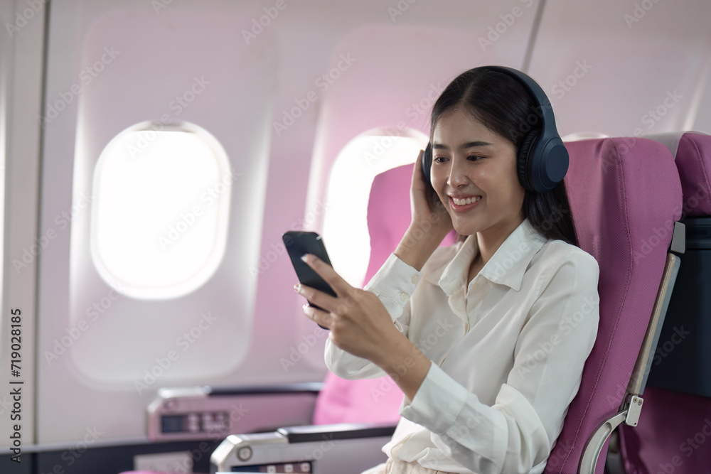 Cheerful Asian woman passenger in headphone watching online movie during intercontinental flight in cabin of aircraft, happy traveler using wifi connection on board Cheerful Asian woman passenger in headphone watching online movie during intercontinental flight in cabin of aircraft, happy traveler using wifi connection on board