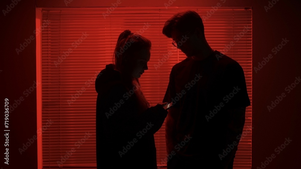Portrait of male in dark room. Handsome man near window, red neon light shines behind jalousie, girl next to man checks smartphone.