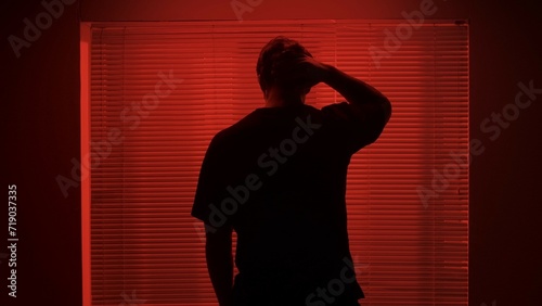 Portrait of male in dark room. Handsome man near window, red neon light shines behind jalousie, guy facing window touching his hair back shot.