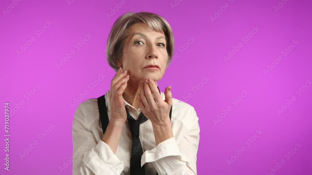 A senior woman is delicately touching her cheeks and looking into the camera, illustrating a mindful skincare routine on a vivid purple backdrop. A senior woman is delicately touching her cheeks and looking into the camera, illustrating a mindful skincare routine on a vivid purple backdrop.