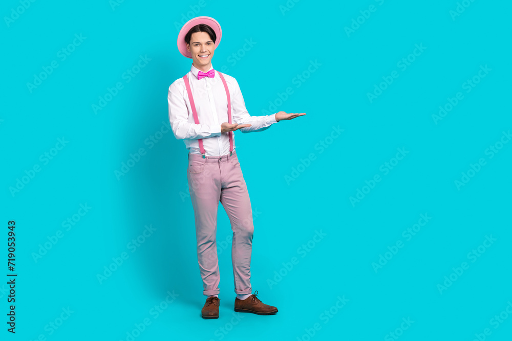 Full length photo of pleasant man dressed cap pants on suspenders palms demonstrate product empty space isolated on blue color background Full length photo of pleasant man dressed cap pants on suspenders palms demonstrate product empty space isolated on blue color background