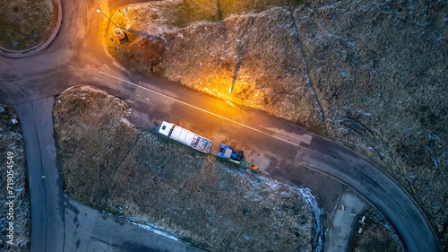 This aerial image captures a lone truck as it traverses a winding road at dusk. The scene is illuminated by a warm streetlight that casts a golden hue on the tarmac, contrasting with the cool tones of