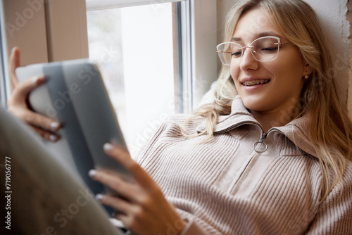 Young woman with braces wearing beige sweater with glasses, smiling with reading tablet by window. Smiling patient. Concept of beauty and medicine, dental care, malocclusion, orthodontic health. photo