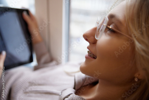 Close up photo of young woman with braces wearing beige sweater with glasses watching movies in tablet by window. Concept of beauty and medicine, dental care, malocclusion, orthodontic health.