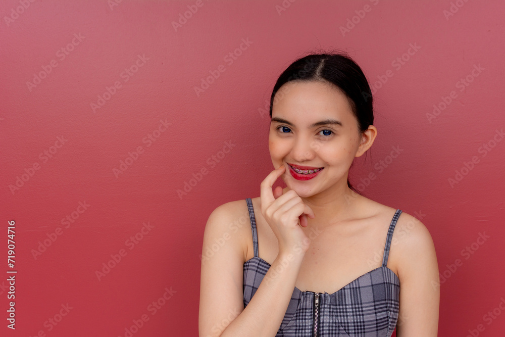Portrait of a cheerful young Asian lady with braces, wearing a plaid dress and posing against a red backdrop. Portrait of a cheerful young Asian lady with braces, wearing a plaid dress and posing against a red backdrop.