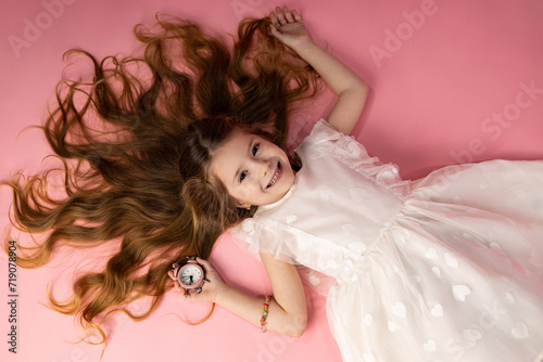 A very wonderful and cute little girl who is lying on a pink background with her hair loose and laid very nicely, brown hair and very fluffy and shiny, the little girl is holding a pink watch in her