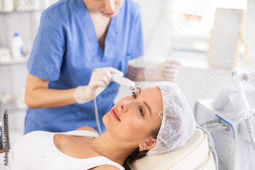 Young woman cosmetologist performs facial microcurrent procedure with machine to young female client