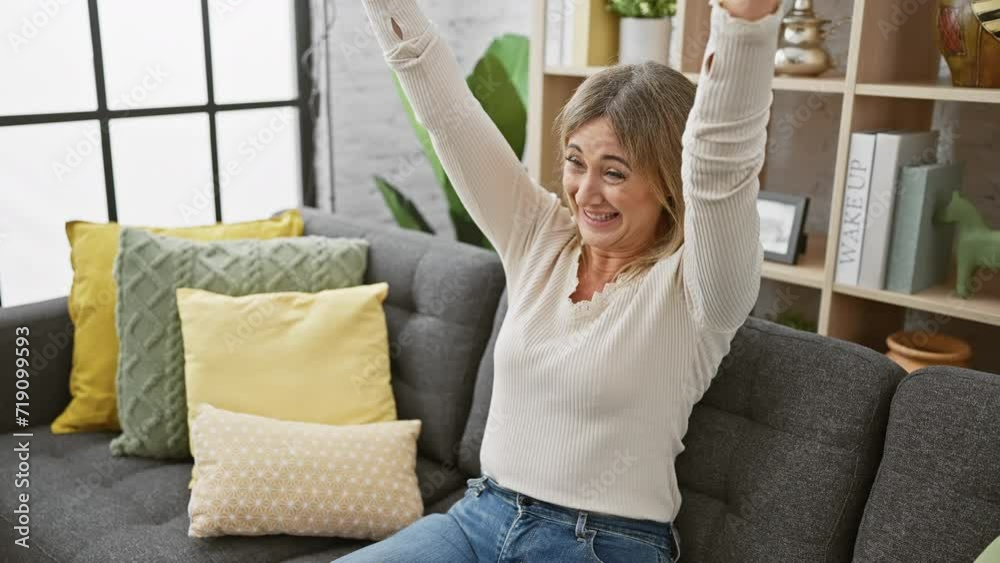 Excited middle-aged blonde sits on sofa, arms raised in a triumphant cheer! proud display of victory and success resonating through her scream in cozy home interior. Excited middle-aged blonde sits on sofa, arms raised in a triumphant cheer! proud display of victory and success resonating through her scream in cozy home interior.