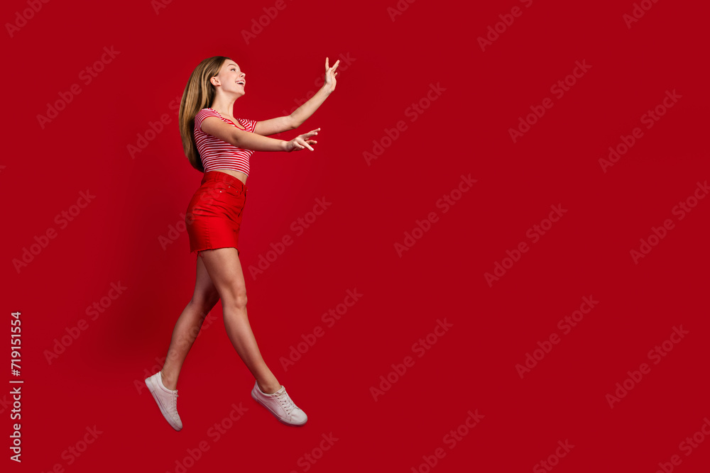 Full length photo of adorable excited girl wear striped top short skirt jumping walking holding arms empty space isolated red color background