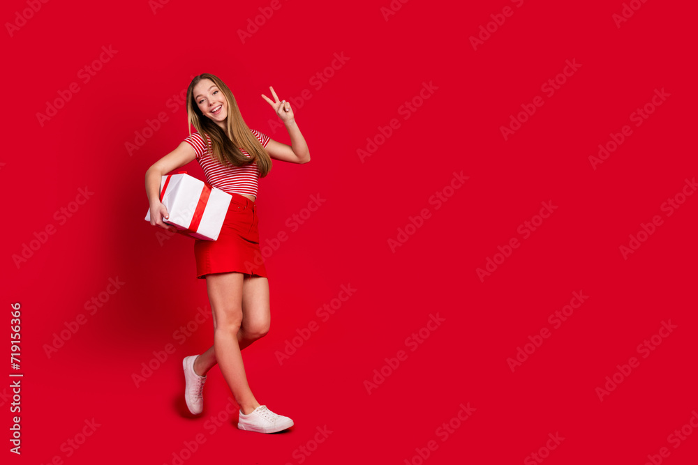 Full length photo of excited funky girl wear striped top short skirt carrying gift boxes showing v-sign empty space isolated red color background Full length photo of excited funky girl wear striped top short skirt carrying gift boxes showing v-sign empty space isolated red color background