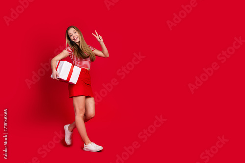 Full length photo of excited funky girl wear striped top short skirt carrying gift boxes showing v-sign empty space isolated red color background