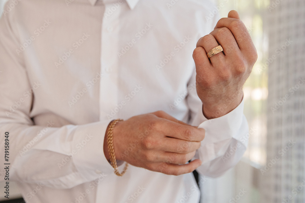 Obraz premium A man with a gold wedding ring and a bracelet fastens the collar of a white shirt, close-up photo