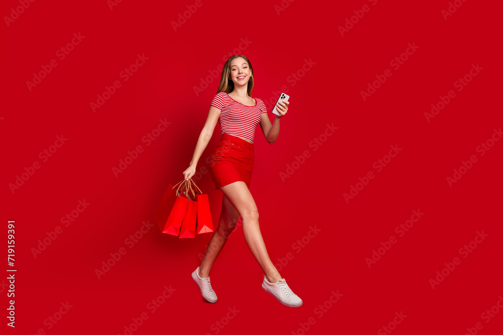 Full length photo of pretty excited girl wear striped top holding bargains shopping modern device isolated red color background Full length photo of pretty excited girl wear striped top holding bargains shopping modern device isolated red color background