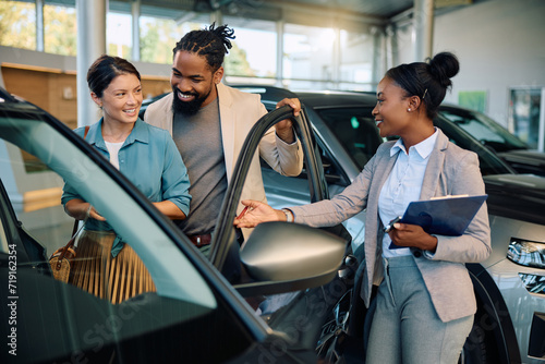 Happy multiracial couple communicating with saleswoman while buying new car in showroom.