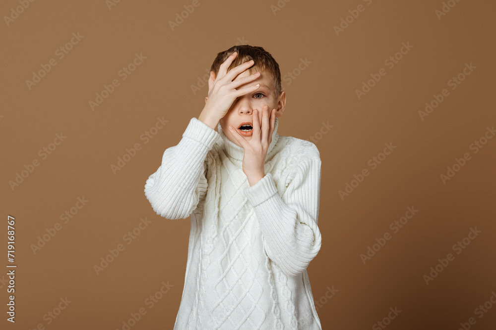 Shocked boy touches face and looks concerned, worried of something, standing over beige background in white knitted sweater Shocked boy touches face and looks concerned, worried of something, standing over beige background in white knitted sweater