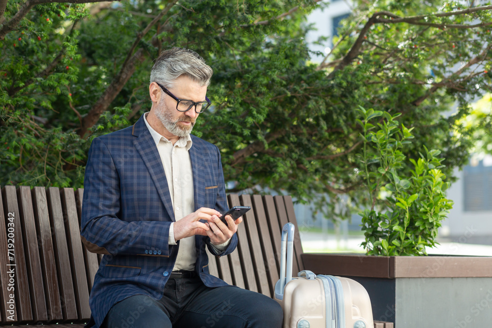 Fototapeta premium Mature man in a business suit using a phone while sitting on a bench outside an office with a suitcase, portraying a professional on the move.