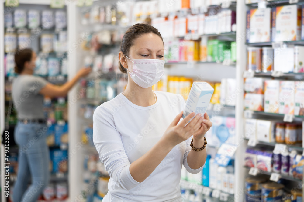 Adult woman in casual clothes with medical mask on her face chooses remedy in pharmacy