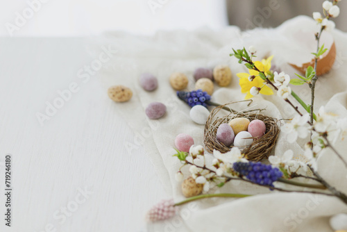 Happy Easter! Stylish easter chocolate eggs in nest, spring flowers, feathers and linen cloth on white rustic wooden table. Easter modern simple banner, space for text. Seasons greetings