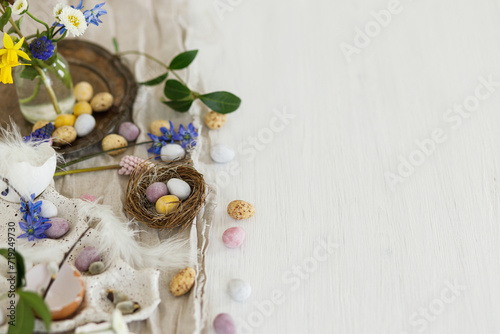 Colorful easter chocolate eggs in nest, spring flowers, feathers and linen cloth on rustic wooden table. Space for text. Easter modern simple decoration. Happy Easter!