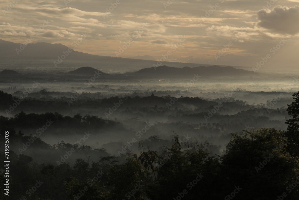 candi borobudur shrouded in mist, with the mountains in the background on a sunny morning.