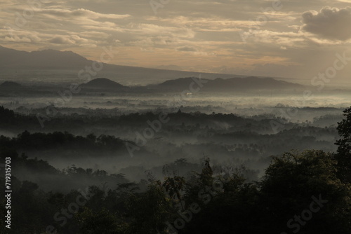 candi borobudur shrouded in mist, with the mountains in the background on a sunny morning.