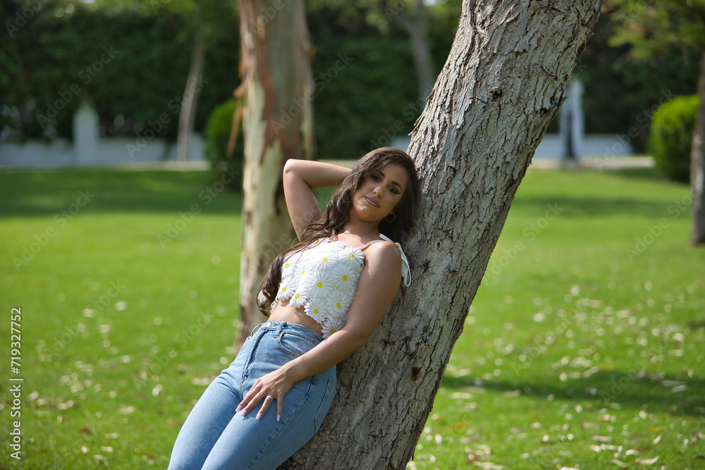 Young woman, Hispanic, beautiful, brunette, with daisy t-shirt and jeans, leaning on a tree trunk, calm and relaxed. Concept of beauty, fashion, trend, nature, spring.