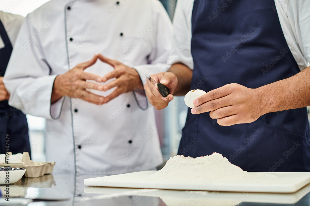 cropped view of young african american man in apron breaking egg next to chief cook, confectionary cropped view of young african american man in apron breaking egg next to chief cook, confectionary