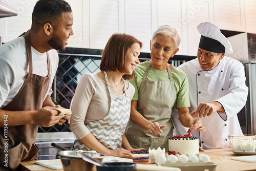 mature woman decorating cake and looking at her jolly diverse friends next to chef, cooking courses photo