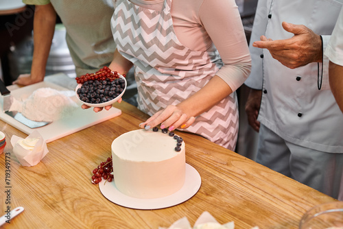 cropped view of young woman decorating cake with berries next to her mature friend and chef