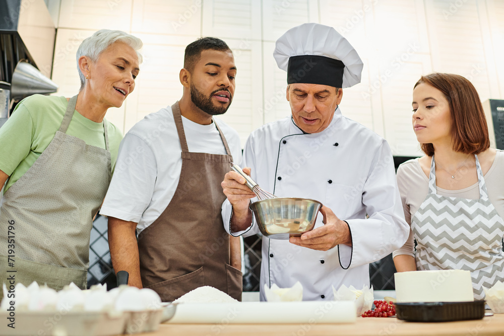 handsome mature chef in white hat showing how to whisk dough to diverse students, cooking courses handsome mature chef in white hat showing how to whisk dough to diverse students, cooking courses