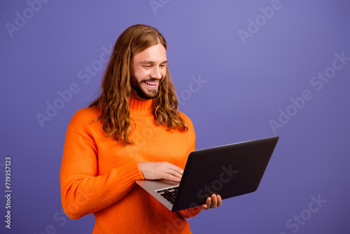 Photo of young happy hipster guy in orange jumper using laptop browsing google analyzing searches isolated on violet color background