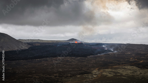 Volcano eruption in Iceland, a fountain of glowing-red lava rising above a vent and a lava flow spreading rapidly downhill, aerial shot.