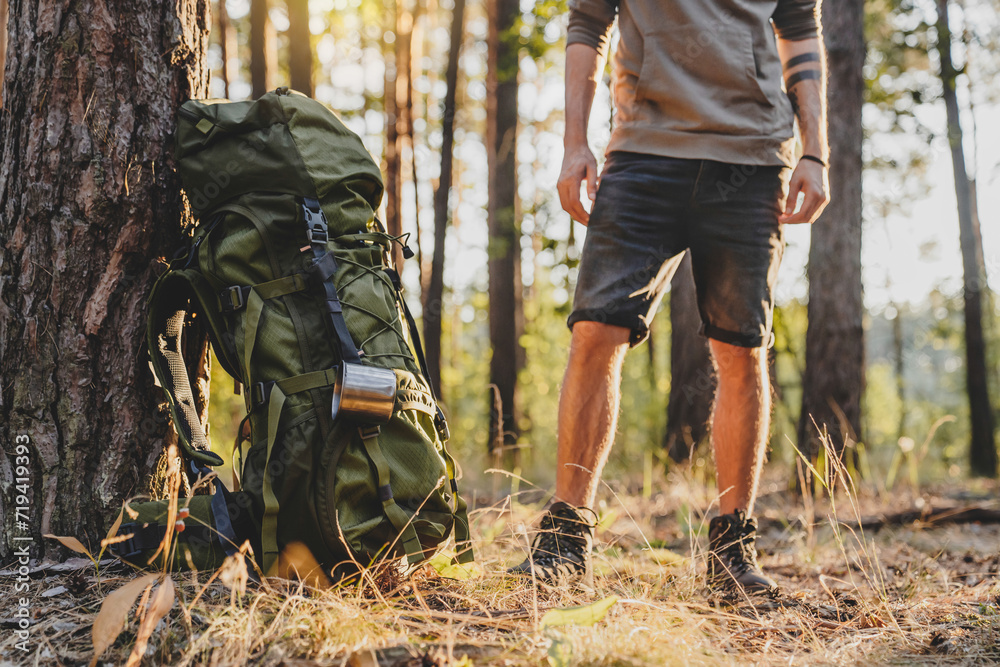 Cropped of male hiker standing near his camping backpack in forest. Away from everything concept. Adventurer explorer traveler in woods in summer