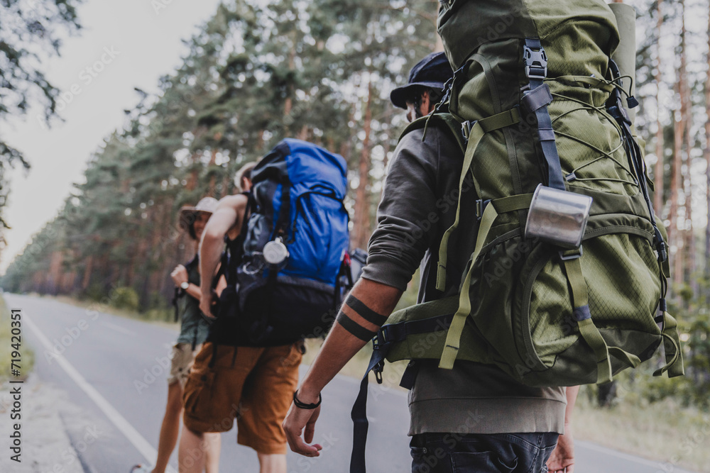 Group of friends traveling with backpacks while walking through forest road. Away from everything concept. Trip voyage to wildlife. Hitchhiking on the road Group of friends traveling with backpacks while walking through forest road. Away from everything concept. Trip voyage to wildlife. Hitchhiking on the road