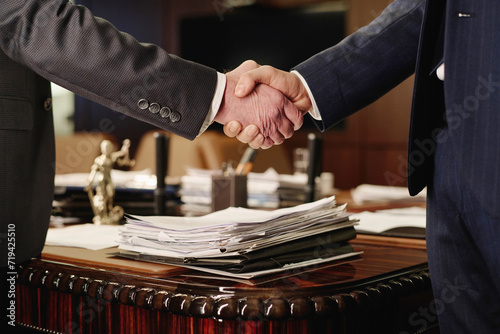 Closeup of two unrecognizable businessmen shaking hands standing next to table with pile of papers